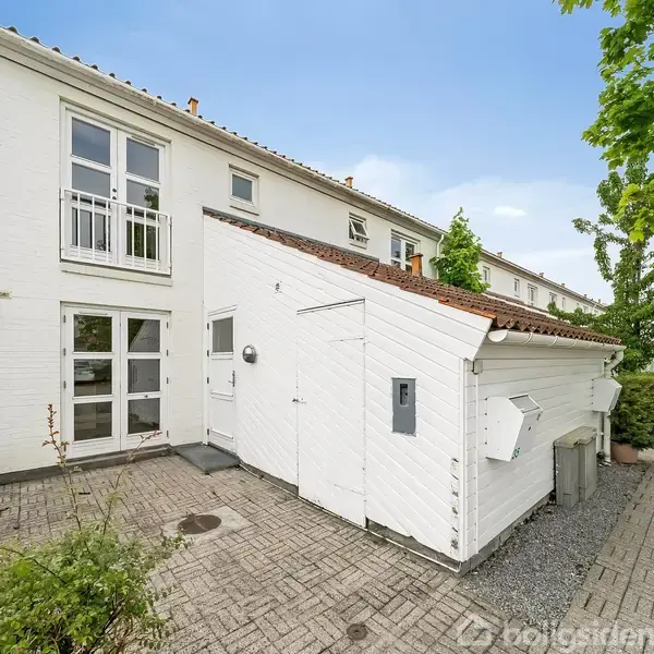 Rækkehus med hvid facade og rødt tegltag. Dets gårdhave har fliser og planter. En gangvej løber langs siden, omkranset af grønne træer.