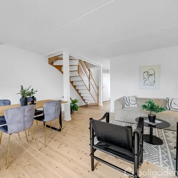 En moderne stue indeholder en lys træspisebord med blå stole til venstre, og en sofa og to sorte lænestole omkring et sofabord til højre. Trappe i baggrunden.