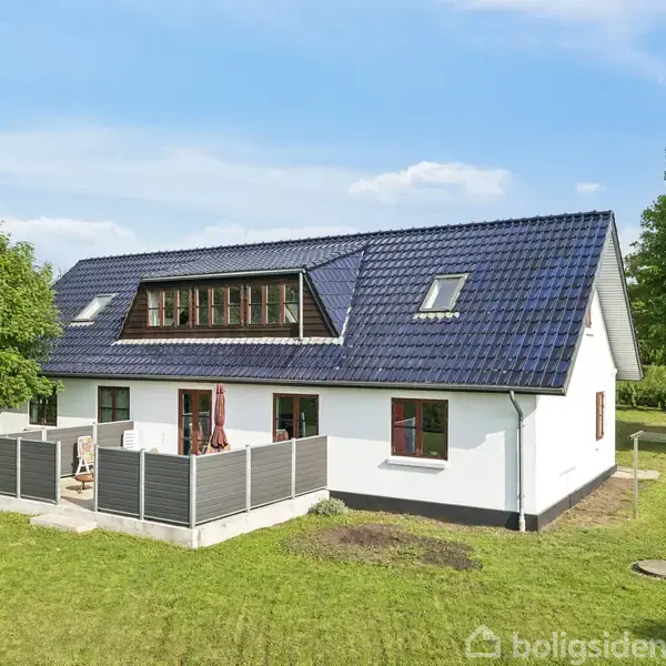 Hvidt hus med blå tagsten og flere vinduer står på en grøn græsplæne omgivet af træer under en blå himmel. Terrassen har møbler og et hegn.