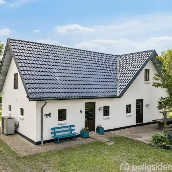 Et hvidt hus med blå tagsten står på en grøn grund. En blå bænk står foran indgangen ved siden af små potteplanter. Nummeret "5" er på væggen.