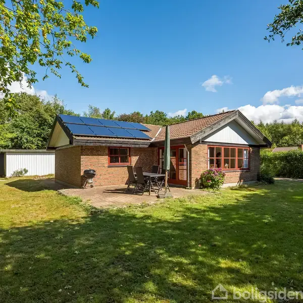 Et murstenshus med solpaneler på taget står i en frodig have. Foran huset er et terrasseområde med havemøbler. I baggrunden er der mange træer og en garage.