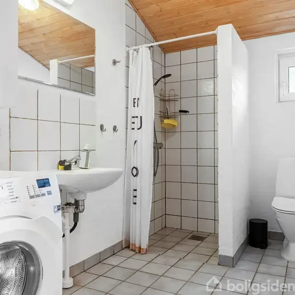 Vaskemaskine og håndvask ved siden af flisebelagt bruseområde. Skillevæg adskiller bruseniche fra toilet. Træloft og lille vindue i hvidt badeværelse med funktionsdygtigt, moderne design.