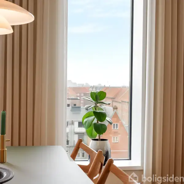 En grøn plante står på et bord foran et stort vindue med udsigt over byens tage. Et tændt lys i en lysestage og en skål med bananer ses på bordet.