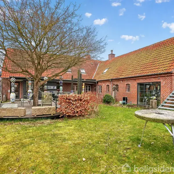 Et rødt murstenshus med rødt tegltag ligger i en have med en terrasse under et træ. Kunstværker pryder terrassen. Til højre ses en trappe, der fører op til døren.