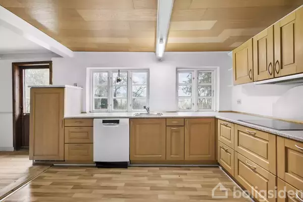 Et køkken med lyse træskabe og en opvaskemaskine i midten. Store vinduer giver udsigt til træer udenfor. Rummet har trægulv og et lyst loft med en lampe.