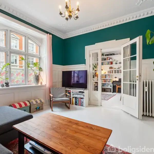 Stue med sofa og træbord, vendt mod et åbent dobbelt glasdør, der fører til et rum med bogreol. Grønlige vægge, hvide paneler og store vinduer med lyserøde gardiner.