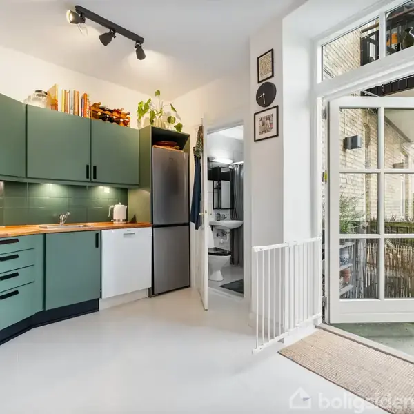 Køkken med grønne skabe og træbordplade, indeholder køleskab og ovn, belyst af spotlys. En åben dør fører til en terrasse. Der er planter og dekorationer på skabene.