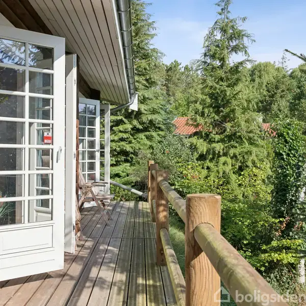 En træterrasse med åbne glasdøre grænser op til et træhus. Terrasse omgivet af frodige træer og tæt skov, hvilket skaber en fredfyldt og naturlig atmosfære.