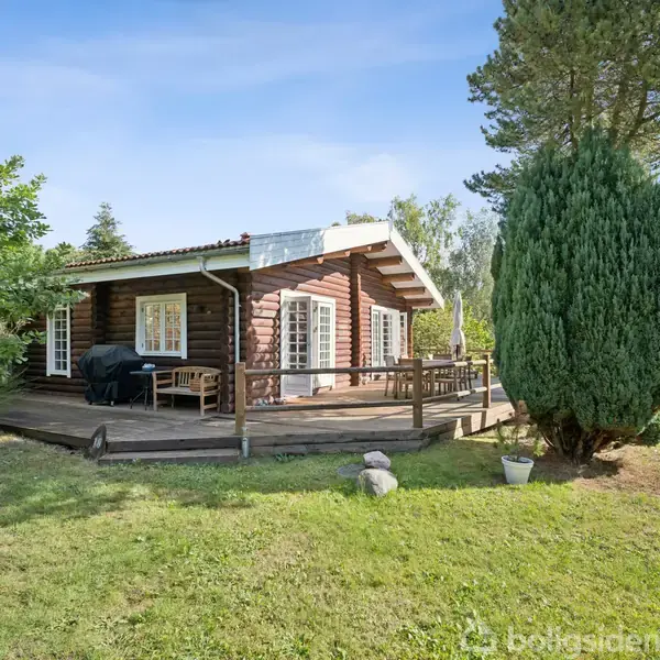 Et træhus står stille på en græsplæne omgivet af frodig skov. Der er en stor terrasse foran huset med udendørs møbler placeret til afslapning.