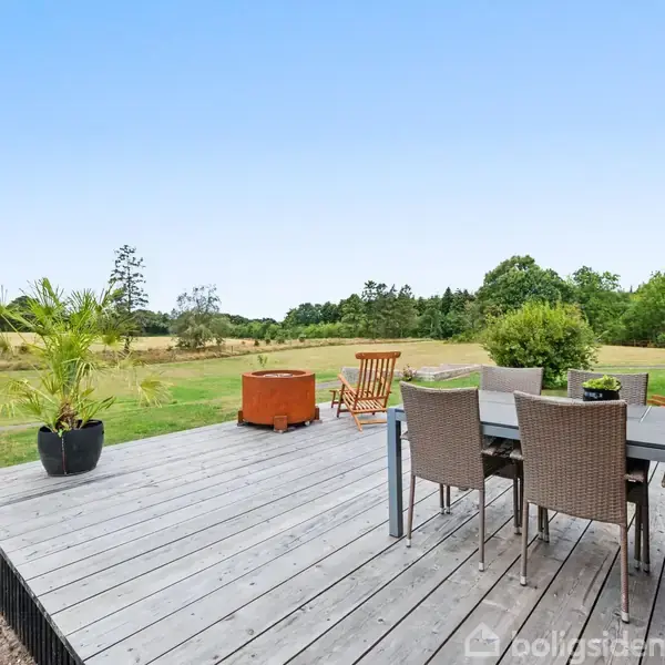 En træterrasse med fem stole og et bord står foran et hus, omgivet af en grøn have med træer i baggrunden under en klar blå himmel.