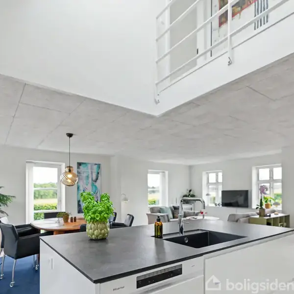 Køkken med køkkenø og vask, udsigt til spiseområde med bord og stole, og stue med sofa og lænestol. Store vinduer giver lysindfald, og der ses en åben førstesal.