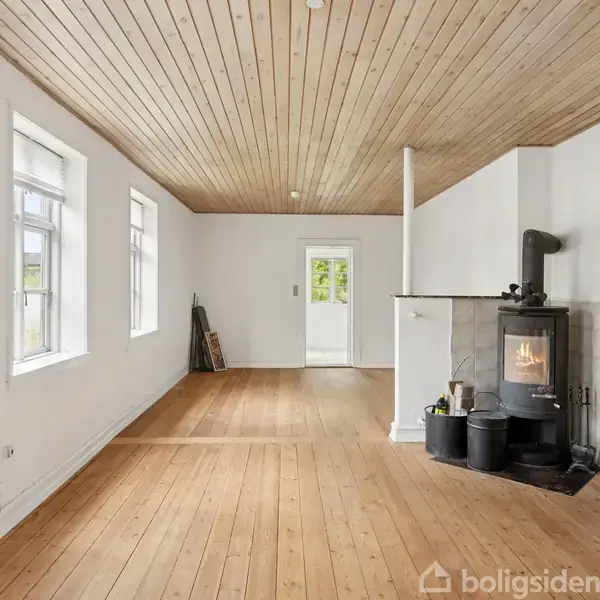 Stue med brændeovn, træloft og gulve, placeret tæt ved væg. Rummet har hvide vægge, flere store vinduer og åbner op mod en lys dør til udendørsområde.