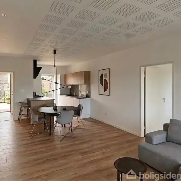 Grå sofa står i en moderne, lyst indrettet stue/køkken område med trægulv. Rundt spisebord og stole nær køkken. Minimalistisk dekoration på væggene, store vinduer til venstre.