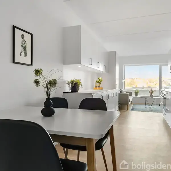 Et moderne køkken med en spiseplads. Sorte stole står ved et hvidt bord med vaser og planter. I baggrunden ses lysindfald fra et vindue ud til en altan.