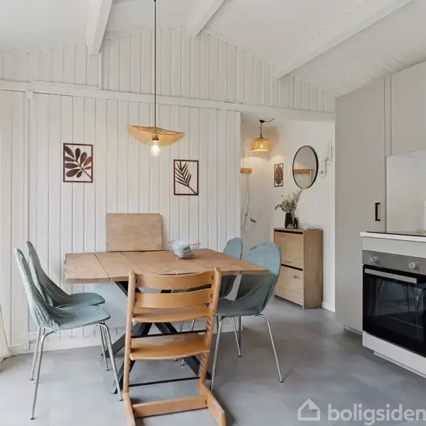 Spisebord med fire stole står i et lyst køkkenalrum med hvid træbeklædning. Vægelamper hænger over bordet, og til højre ses en indbygget ovn og skabe.