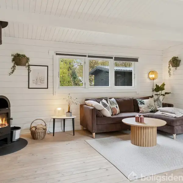 Et moderne stueområde med en brændeovn opvarmer rummet. En hjørnesofa står på et lyst tæppe, omgivet af grønne planter og sofabord. Store vinduer bringer udsigt til udendørs natur.