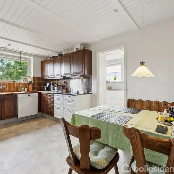 Et køkkenbord med stole er dækket med en dug og skriftmaterialer, placeret nær et stort vindue. Køkkenet har træskabe og en opvaskemaskine, med lyse vægge og loft.