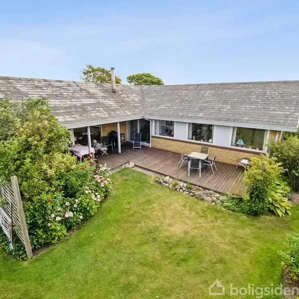 En enetages hus med fladt tag og stor træterrasse. Huset omgives af en velplejet have med grønne plæner og buske, og der er havemøbler på terrassen.