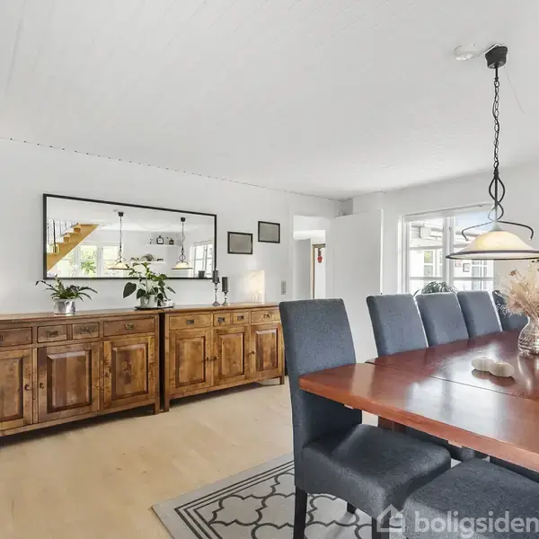 Spisebord med mørke stole står på gulvtæppe; over bordet hænger to lamper. Træskænk med planter og spejl i baggrunden. Lyse vægge og gulv i spisestue.
