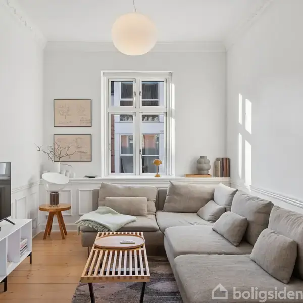 En stor, beige hjørnesofa står i en lys stue med trægulv. En fladskærms-TV er placeret på et hvidt skab. En rund lampe hænger over centrum. Vinduet giver naturligt lys.