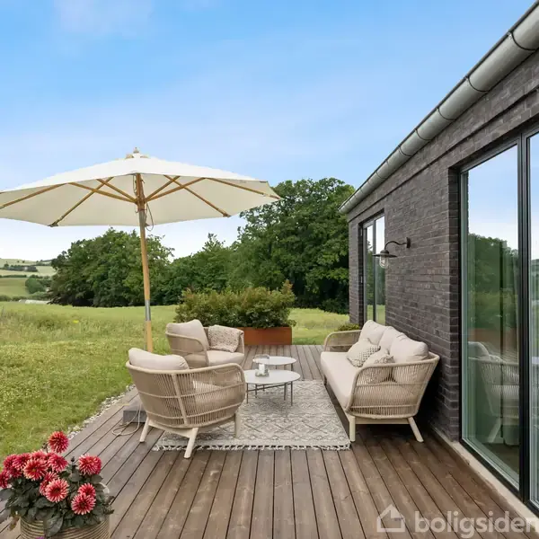 Terrasse med kurvemøbler og parasol står på trædæk, omgivet af frodige marker og træer, ved siden af et moderne hus med store glasvinduer. Farverige blomster pryder forgrunden.