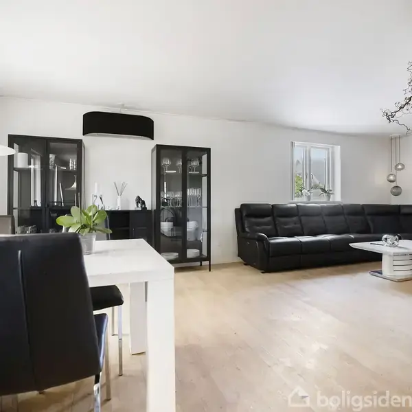 En stor sort lædersofa står langs en væg med et vindue, ledsaget af et hvidt sofabord. Et moderne spisebord med stole ses i forgrunden i et lyst, minimalistisk rum.