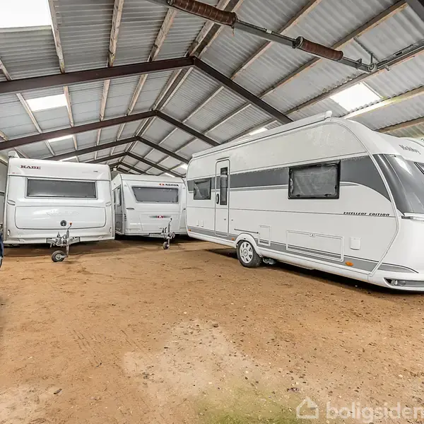 Campingvogne står parkeret i en stor, overdækket lade på jordgulv. Metalstrukturen understøtter taget. Vogne er arrangeret langs siden, med en overdækket bil delvist synlig til venstre.