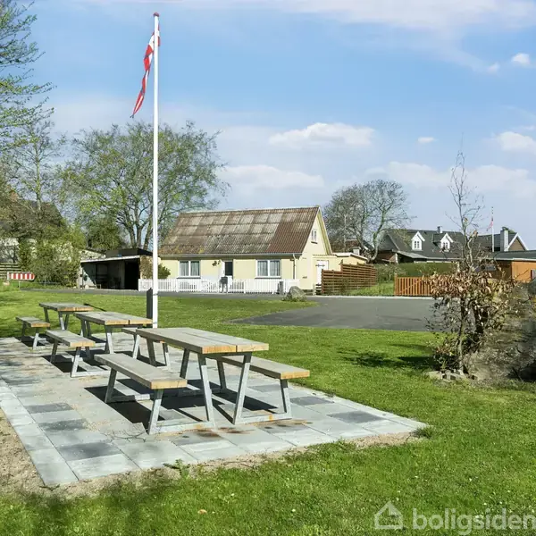 Et picnicbord står på flisebelagt område. Ved siden af står en flagstang med Dannebrog. I baggrunden ses en lys bygning og grønne træer under en blå himmel.