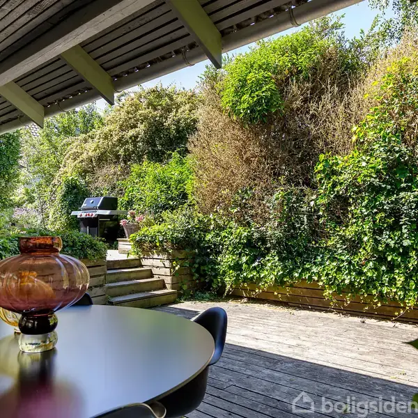 Bord med farvede glasvaser står stille på en terrassedækning med udsigt mod en have fyldt med grønne planter. En grill ses ved trappen under halvtaget.