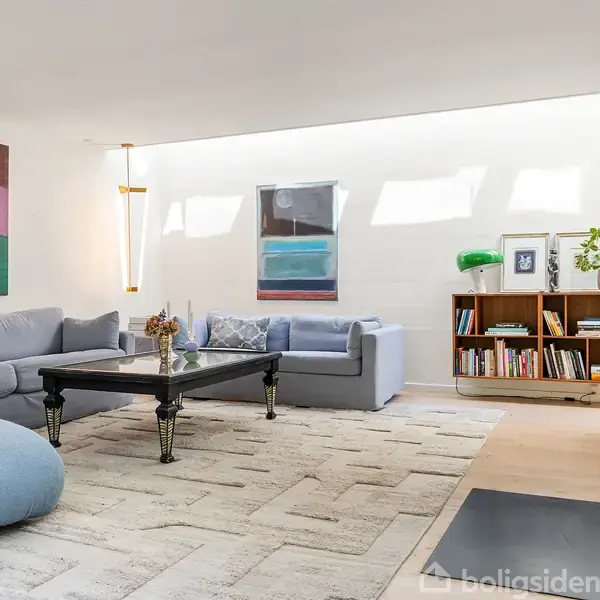 Stue med blå sofaer, et mørkt sofabord med blomster, og bogreol fyldt med bøger. Væggene har moderne kunstværker. Rummet er lyst med naturligt lys og indretning i moderne stil.