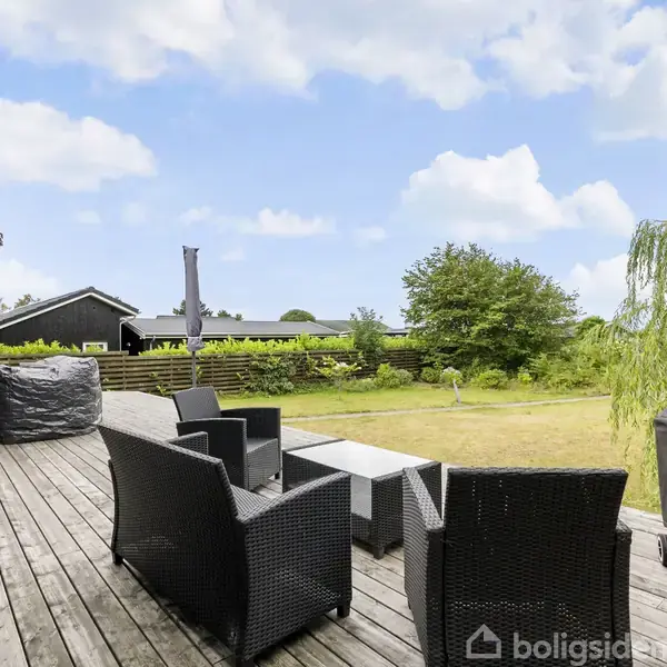 Terrasse med sorte kurvemøbler står ubevægelige, omkranset af en åben have med græsplæne og buske under en klar blå himmel. En grill er placeret til højre.