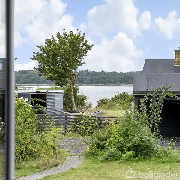 Et træ står ved siden af en sort bygning med en sø i baggrunden. Landskabet er frodigt med buske og træer, omgivet af et roligt vandområde.