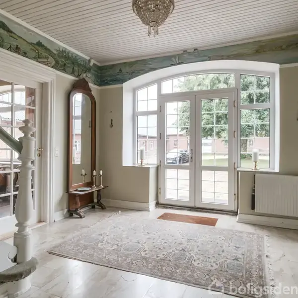 Entré med trappe, hvor lysekronen hænger i loftet. Store buede vinduer og glasdøre åbner mod en udendørs gård. Et tæppe ligger på gulvet, og et spejl står ved væggen.