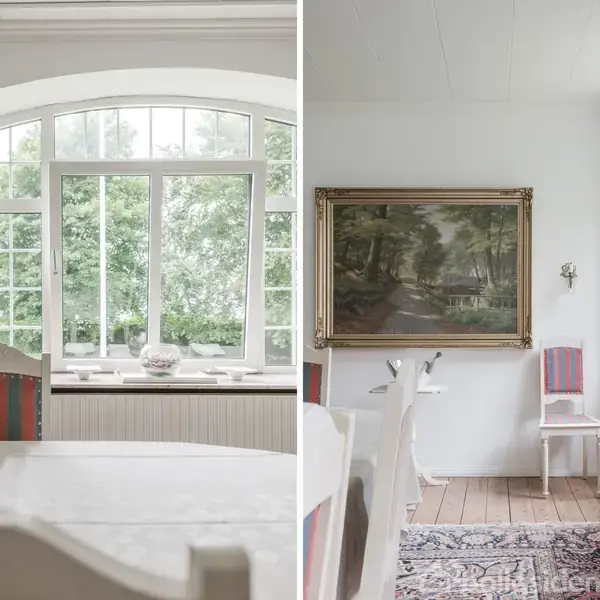 Der er et lyst rum med et stort panoramavindue, der viser grønne træer udenfor. En hvid stol og et maleri af en skovlandskab hænger på væggen ved siden af.