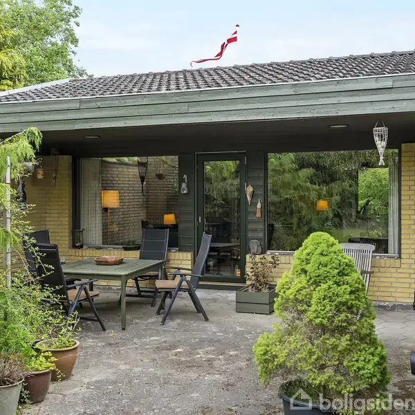 Et hus har en terrasse med et bord og stole. Planter omgiver terrassen, og en lille flagstang med et flag ses på hustaget.