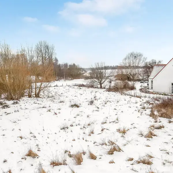 Et hus med rødt tag og hvide vægge står stille i en snedækket landskab. Omgivet af nøgne træer og en klar blå himmel, ser det idylisk og fredfyldt ud.