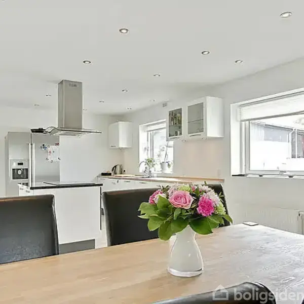 En lys og moderne køkken-spisestue med hvidt interiør, sorte stole og et træbord med en vase med lyserøde blomster. Store vinduer giver godt lysindfald.