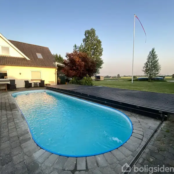 En aflang swimmingpool med blåt vand er omgivet af fliser. Ved siden af er der et hus med terrassemøbler. I baggrunden ses en stor have og en flagstang.