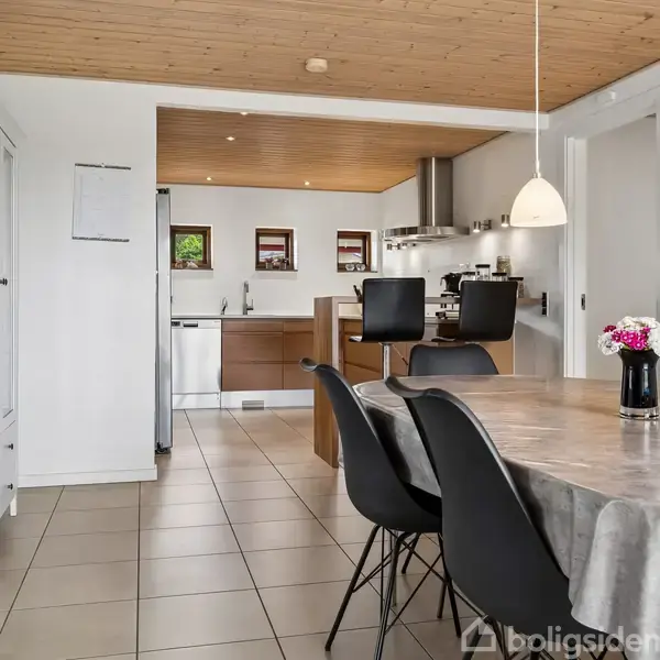 Spisebord med sort stole står i et lyst køkken/alrum. Bordet er dækket med dug og blomster i vase. Loftet er i træ, og køkkenet har moderne indretning med to pendellamper.