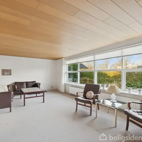 Stue med brune lædermøbler i afslappet opstilling ved siden af et glasbord med lamper. Store vinduer trækker naturligt lys ind, og kunst pryder hvide vægge.