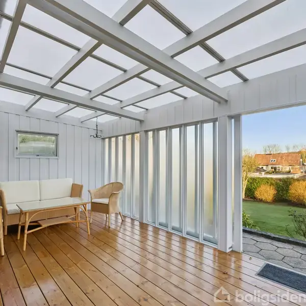 En lys, overdækket terrasse med træplanker og hvide vægge indeholder en sofa og to kurvestole. Den åbne side viser en velplejet have og blå himmel i baggrunden.