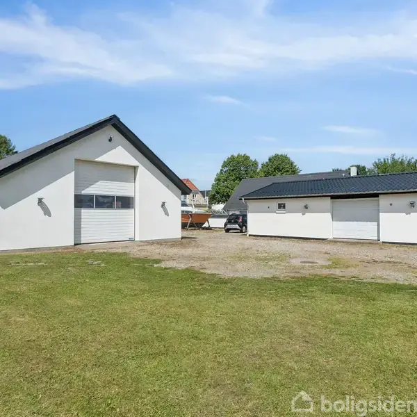 To hvide garageskure med sorte tage står på en gruset gårdsplads. Grønt græs omkranser bygningerne, mens træer og blå himmel danner baggrund.