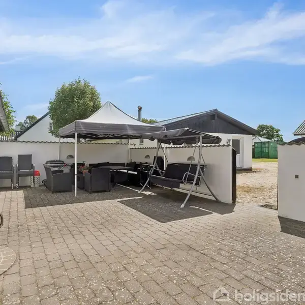 En overdækket terrasse med sorte møbler og en hængekøje står i gården mellem hvide bygninger. Terrassen har to markiser. Møblerne er placeret på en brostensbelagt overflade.