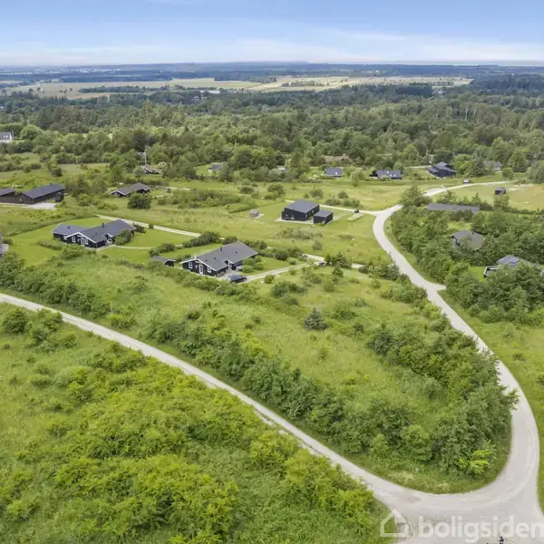 Landskab med flere huse ligger spredt på grønne bakker, forbundet af snoede grusveje. Omkringliggende skove og åbne marker strækker sig mod horisonten under en klar himmel.