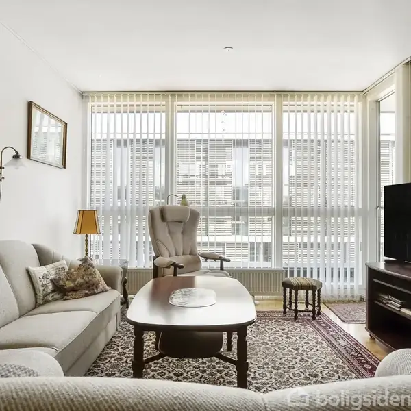 Stue med beige sofa, sofabord og tæppe på gulvet. En stor tv-skærm står på et møbel til højre. Store vinduer med persienner fylder bagvæggen. Væggene har kunstværker.