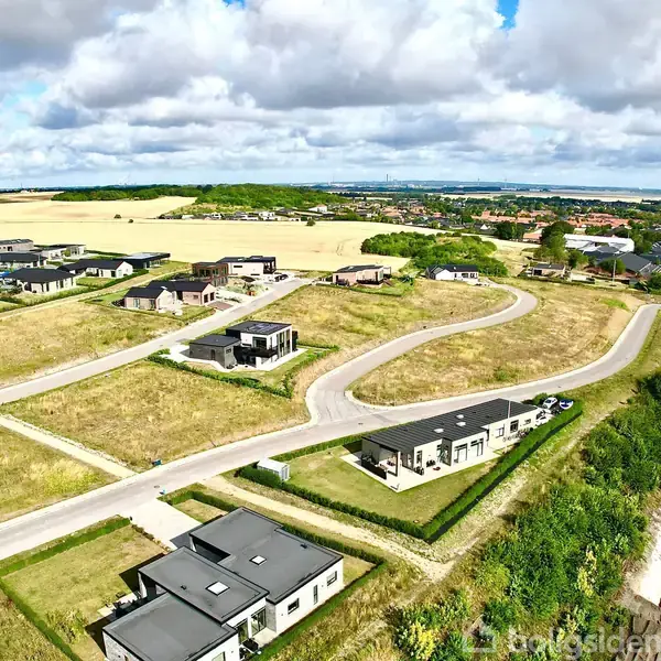 Nyligt byggede enkeltfamiliehuse står på åbne grusveje i en udviklingszone omgivet af åbne marker og grønt landskab under en overskyet himmel. Boliger spredt med regelmæssig afstand.