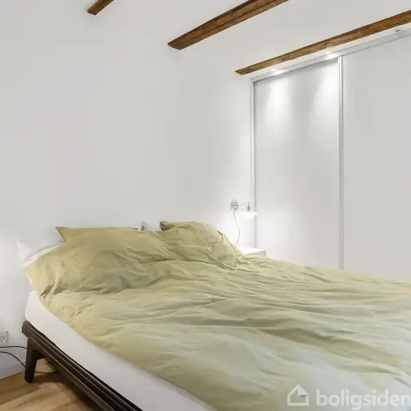 Seng med grønt sengetæppe står i et lyst soveværelse. To natborde med lamper er ved siden af sengen. Væggene har indbyggede skabe, og loftet har synlige bjælker.