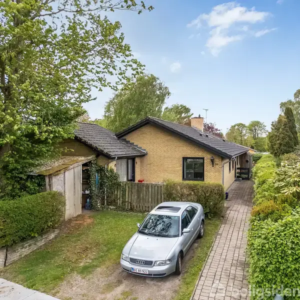 En grå bil står parkeret i en indkørsel ved siden af et gult murstenshus. Omkring huset er grønne træer og buske.