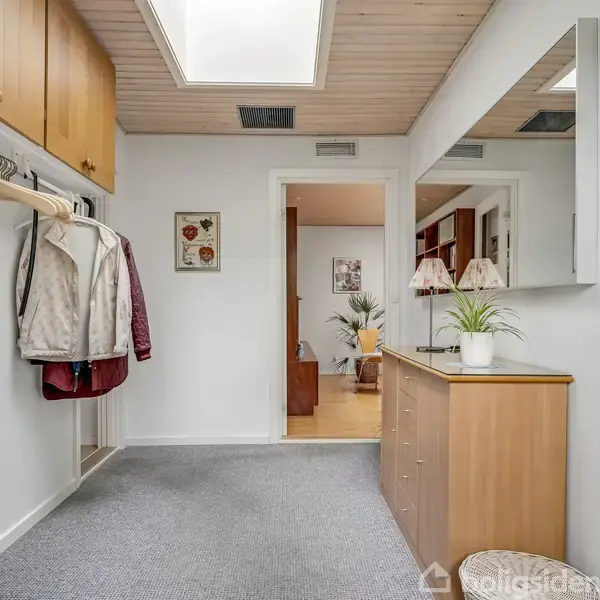 Jakker hænger på en bøjle i en lys gang med tæppe og træloft. Skab med spejl over står til højre. En plante og lampe er på skabet.