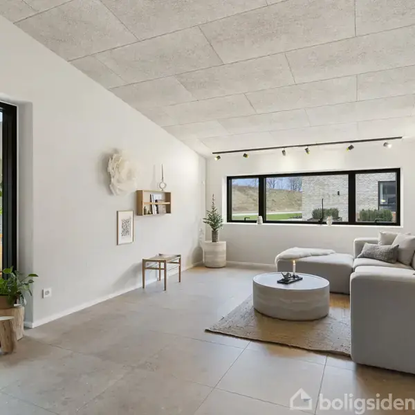 Stue med grå sofa, rundt sofabord og vægkunst. Store vinduer slipper lys ind. Moderat indretning med planter og hylder. Træstubbe bruges som dekoration nær glasdør til udendørsområde.